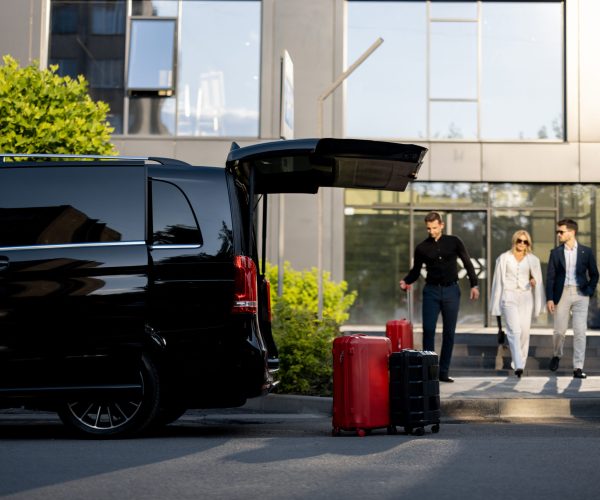 Driver or concierge helps a business couple carry their suitcases to minivan taxi from hotel. Concept of business travel and transportation service